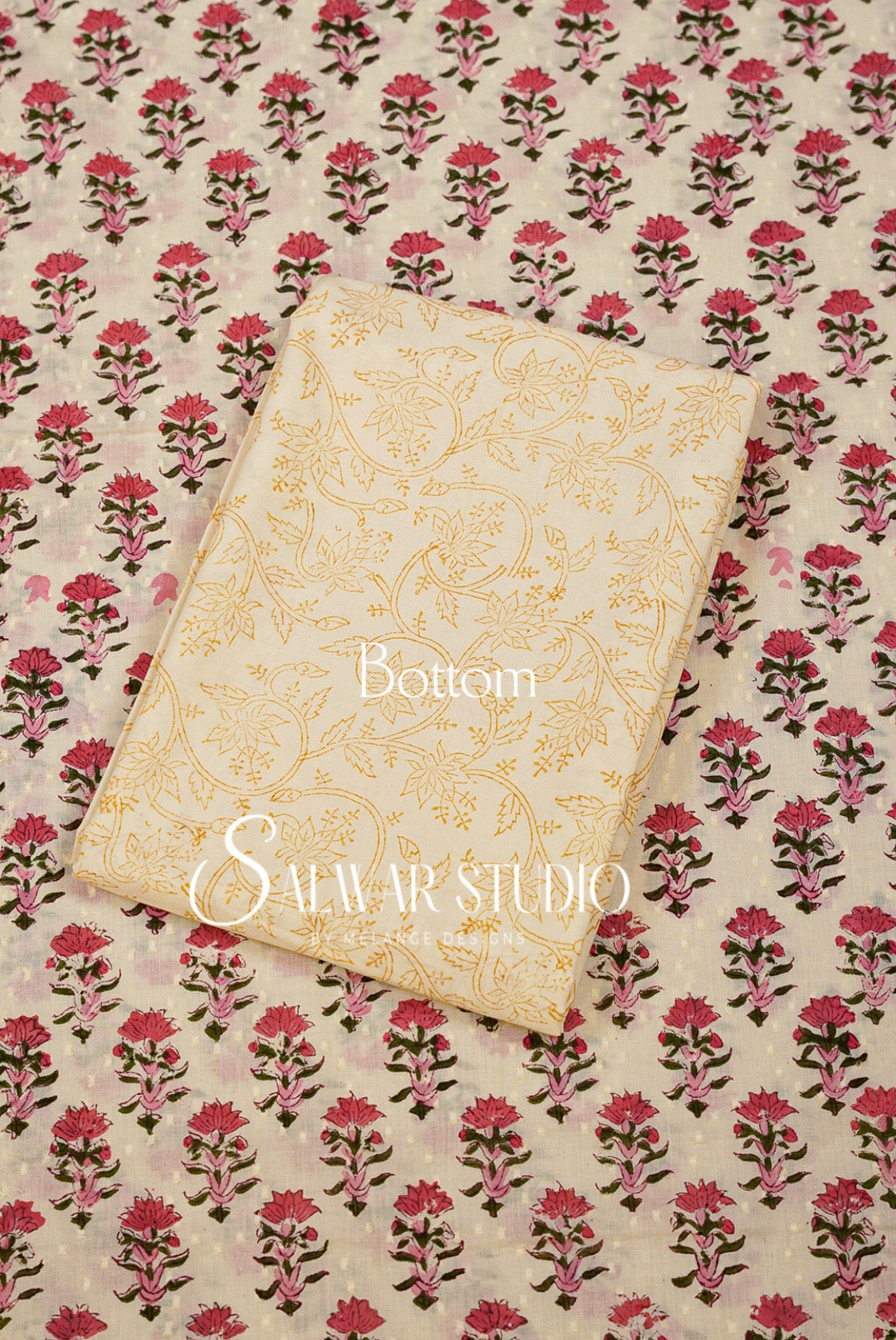 A folded cream-colored fabric piece with gold floral patterns lies on top of a larger matching fabric sheet featuring a repeating pink and green block print on a cream background.