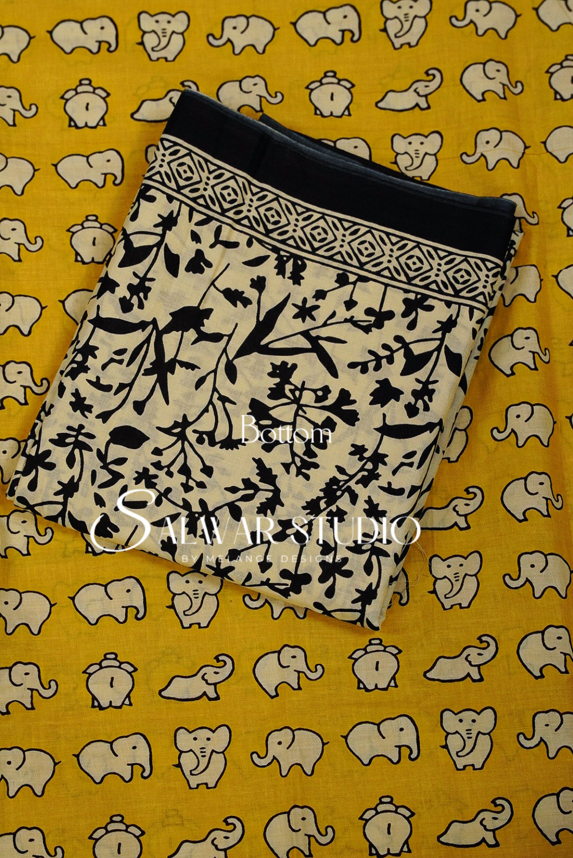 A folded piece of beige fabric with a black floral and bird print, laid flat on a yellow fabric background patterned with beige elephants.