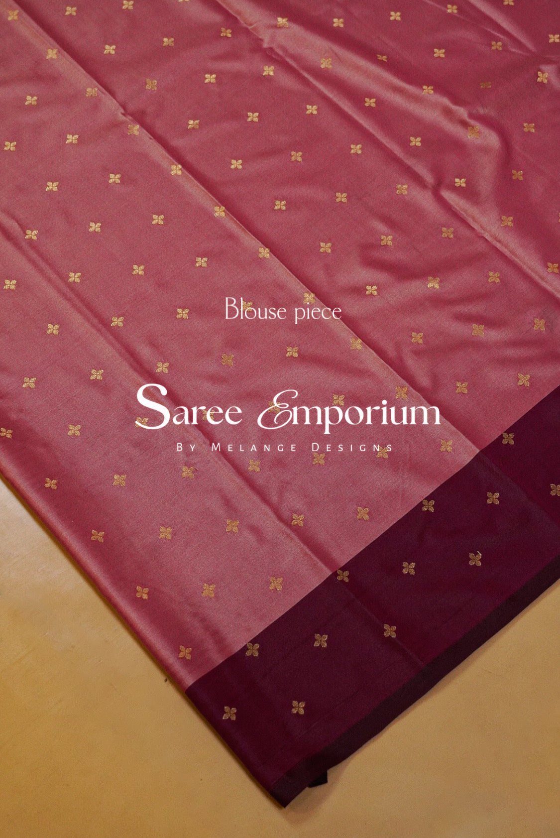 A close-up of a pinkish-red saree fabric with a border in a darker maroon shade, decorated with small gold motifs.