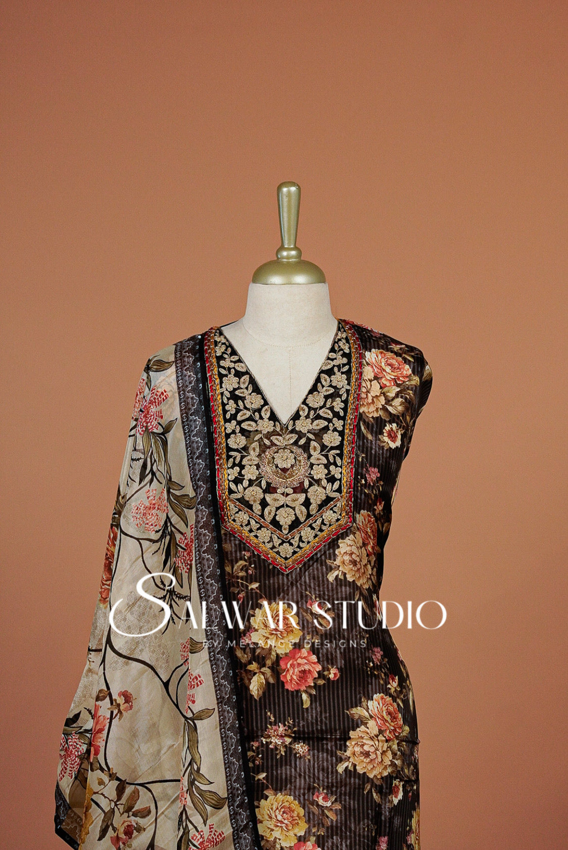 A traditional South Asian salwar suit set displayed on a mannequin against a solid terracotta background. The set includes a black heavily embroidered kurta with floral prints, a beige printed dupatta, and a matching black embroidered border.