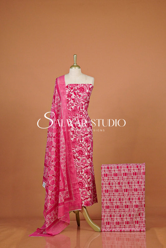 A pink printed salwar suit set  with a matching fabric swatch box beside it. The set includes a printed salwar, a matching dupatta, and a folded fabric sample.
