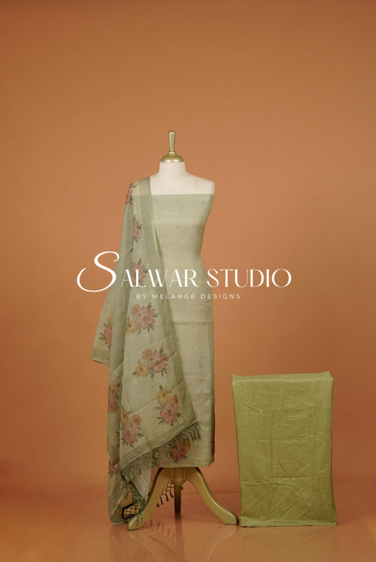 A sage green salwar suit set featuring a printed dupatta with pink and yellow floral motifs, a matching green skirt, and a folded green fabric piece. 