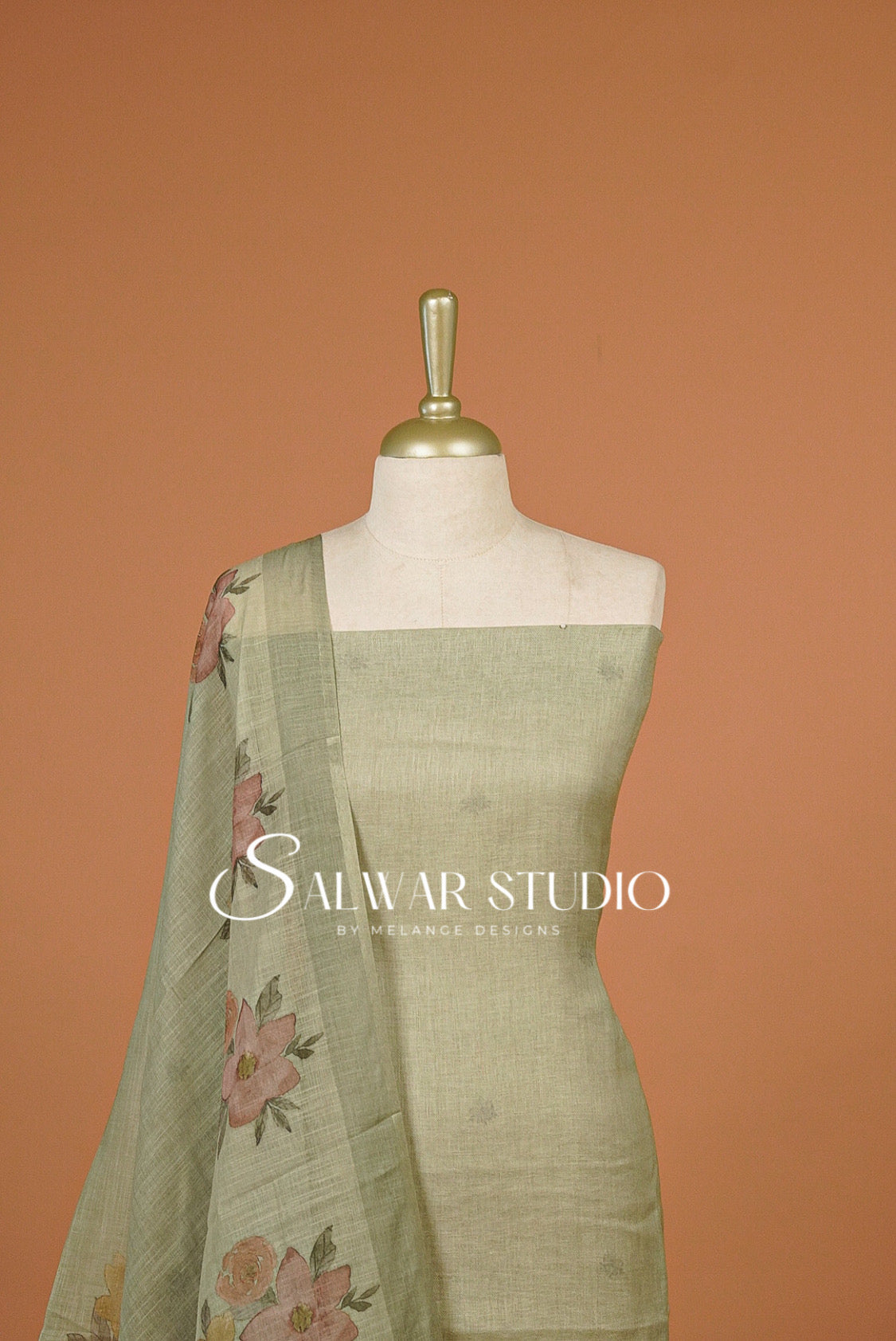 A sage green linen salwar suit set .The set includes a straight-front kurta, matching trousers, and a draped dupatta featuring a pink and green floral border print.