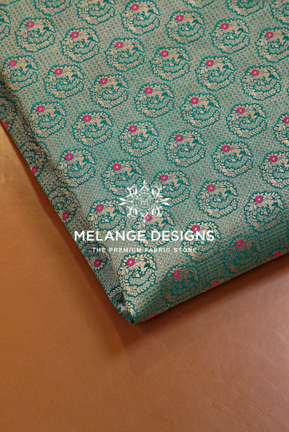 A close-up of folded teal Banarasi Silk Brocade Banarasi Silk Brocade Fabric with an allover floral and ornamental woven pattern in pink, gold, and cream, placed on a warm brown surface. 