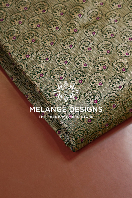 A folded piece of Banarasi Silk Brocade Banarasi Silk Brocade Fabric with an intricate repeating pattern of circular motifs in olive green, gold, and pink, displayed on a warm terracotta surface.