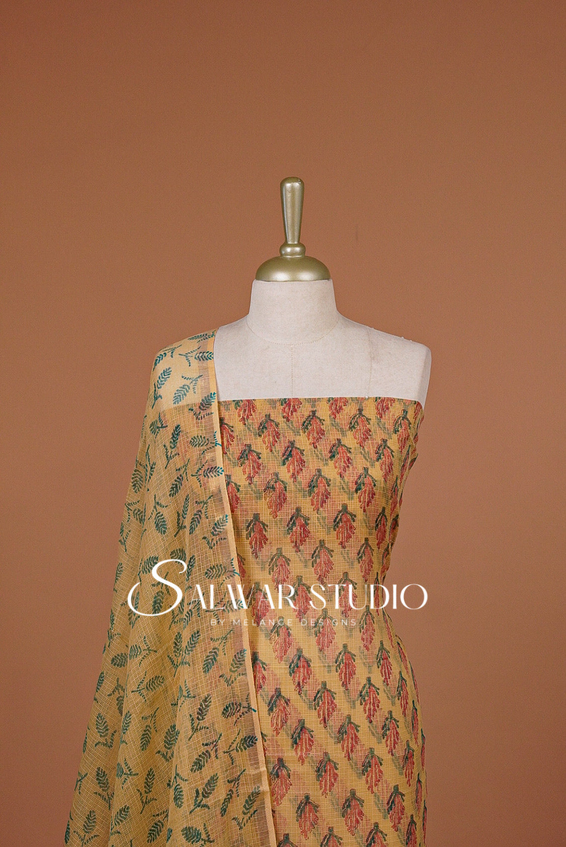 A traditional Indian salwar suit set displayed on a mannequin torso against a solid terracotta background. The set features a mustard yellow base with a red and green geometric pattern on the kurta, and a matching printed dupatta draped over the shoulder.
