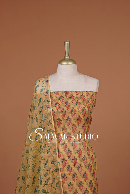 A traditional Indian salwar suit set displayed on a mannequin torso against a solid terracotta background. The set features a mustard yellow base with a red and green geometric pattern on the kurta, and a matching printed dupatta draped over the shoulder.