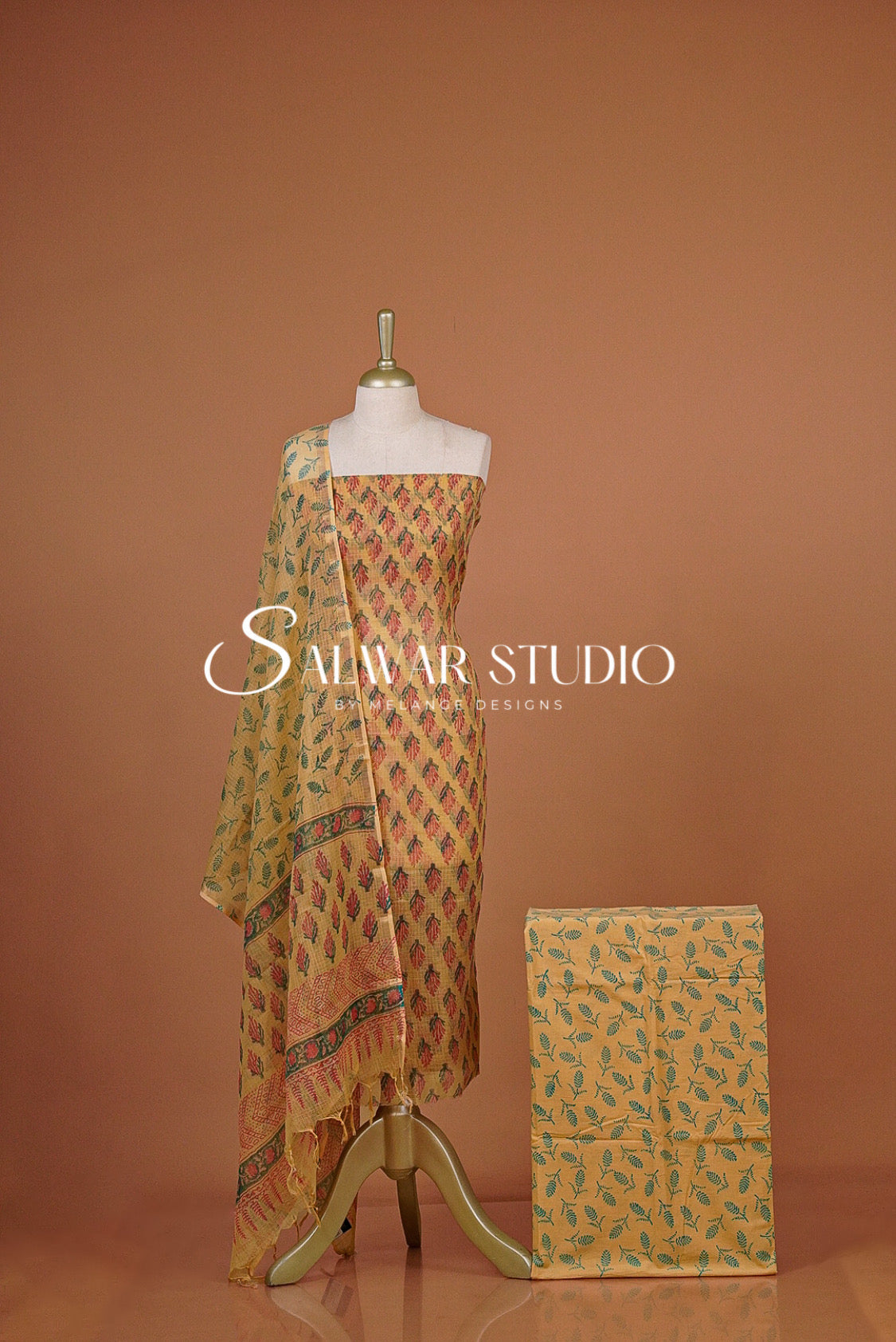 A traditional Indian salwar suit set displayed on a mannequin and folded fabric block against a solid terracotta background. The set features a beige kurta with a multicolor geometric print, a matching dupatta, and coordinating trousers.