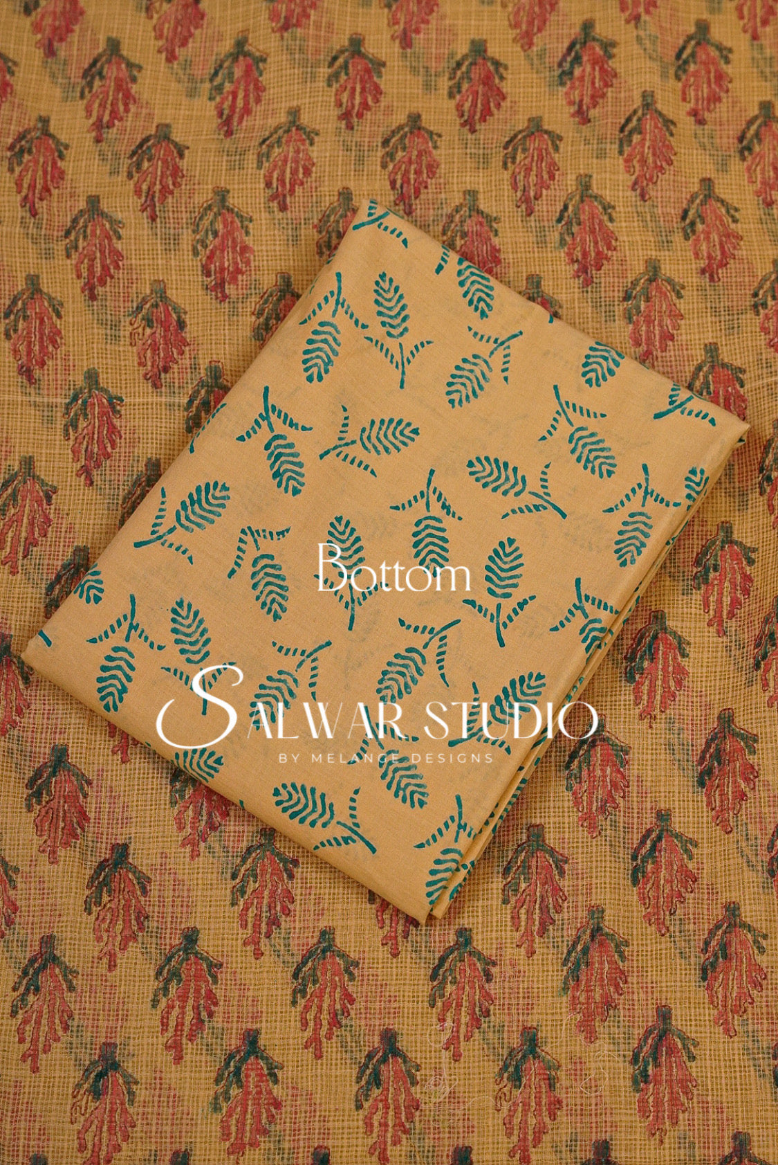 A folded piece of beige fabric with a teal leaf block print pattern, laid diagonally over a larger matching fabric sample showing a red and teal floral block print.