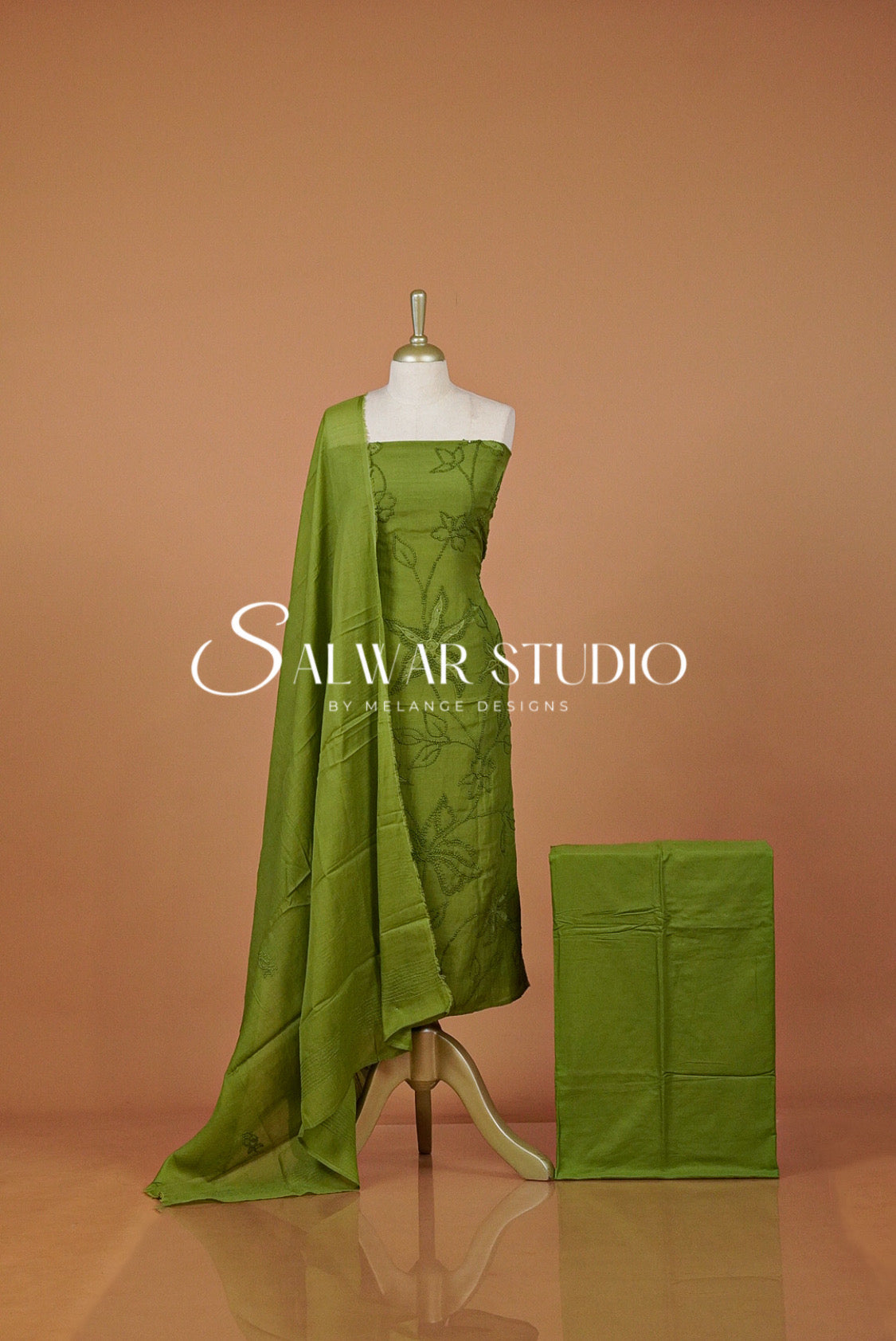 An olive green salwar suit set displayed on a mannequin, featuring a printed kurta, a draped dupatta, and a folded matching fabric piece.