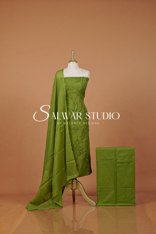 An olive green salwar suit set displayed on a mannequin, featuring a printed kurta, a draped dupatta, and a folded matching fabric piece.
