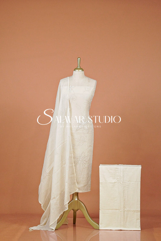 An off-white embroidered salwar suit set displayed on a dress form, featuring a kurta, matching trousers, and a draped dupatta, alongside a folded matching garment .