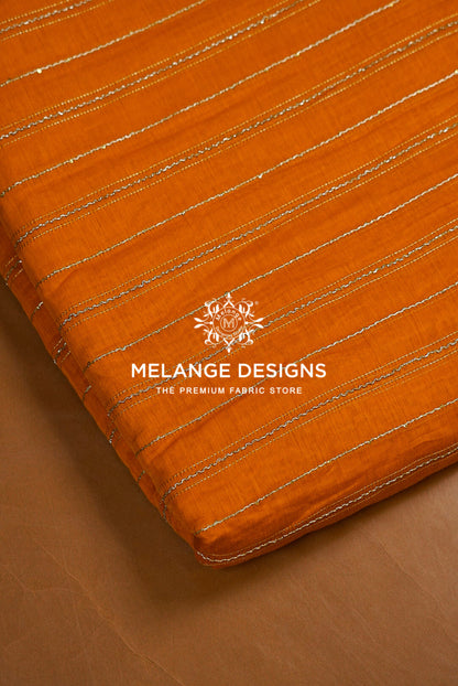 A close-up of an orange Chanderi Silk Embroidery Fabric with thin metallic gold stripes, folded over to show its texture and edge. 