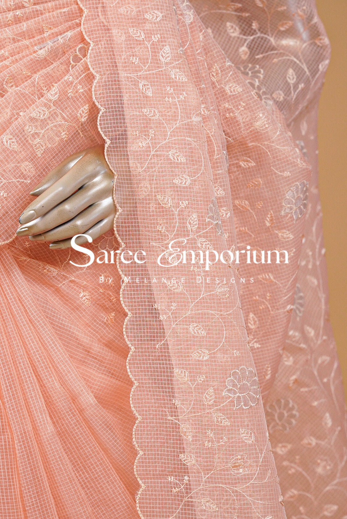 A close-up of a peach-pink saree with a sheer floral embroidered border and scalloped edge, draped over pleated fabric. A metallic hand is visible behind the fabric.