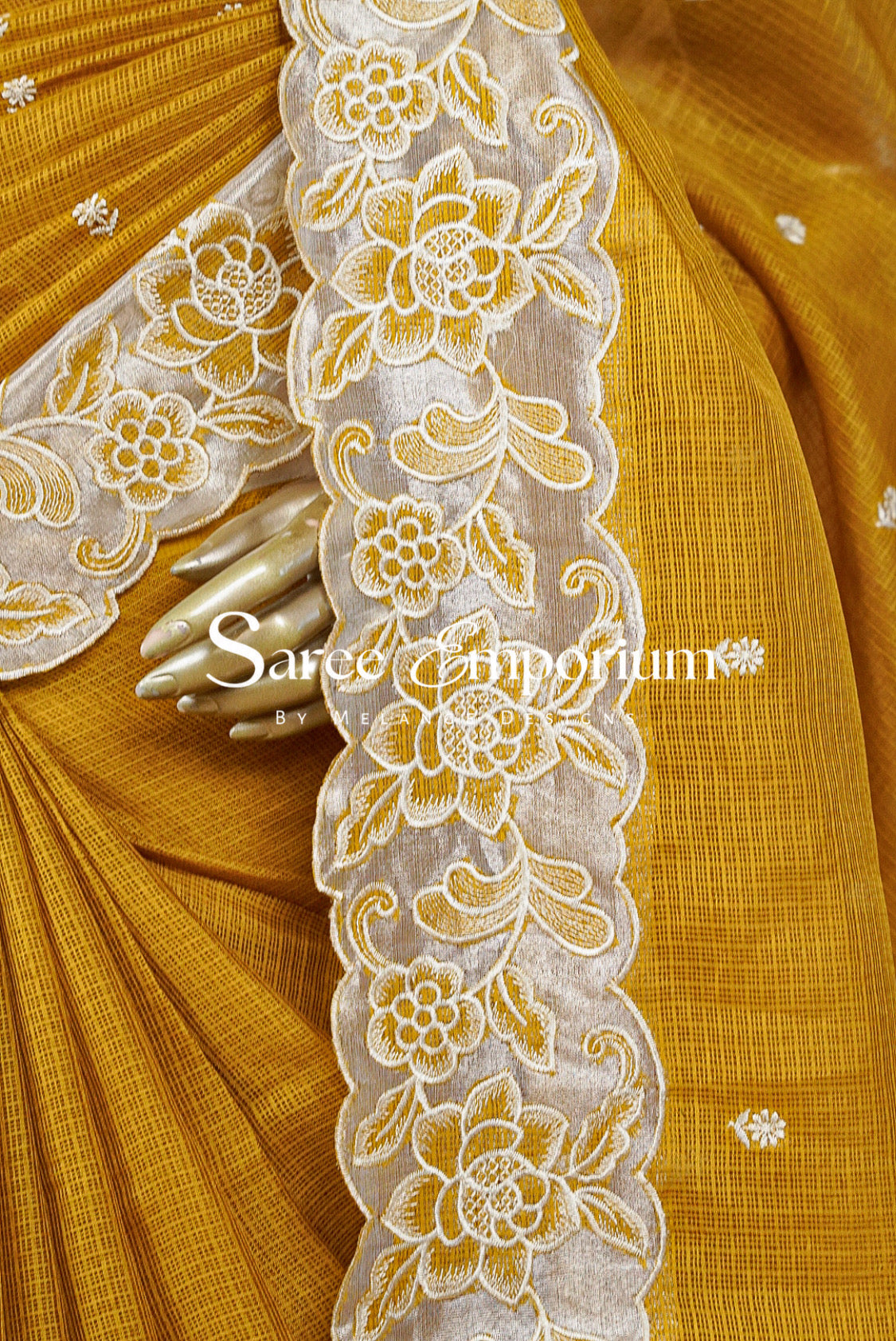 A close-up of a mustard yellow saree with a wide cream lace border featuring embroidered floral patterns. A hand is holding the lace to show its drape and texture.
