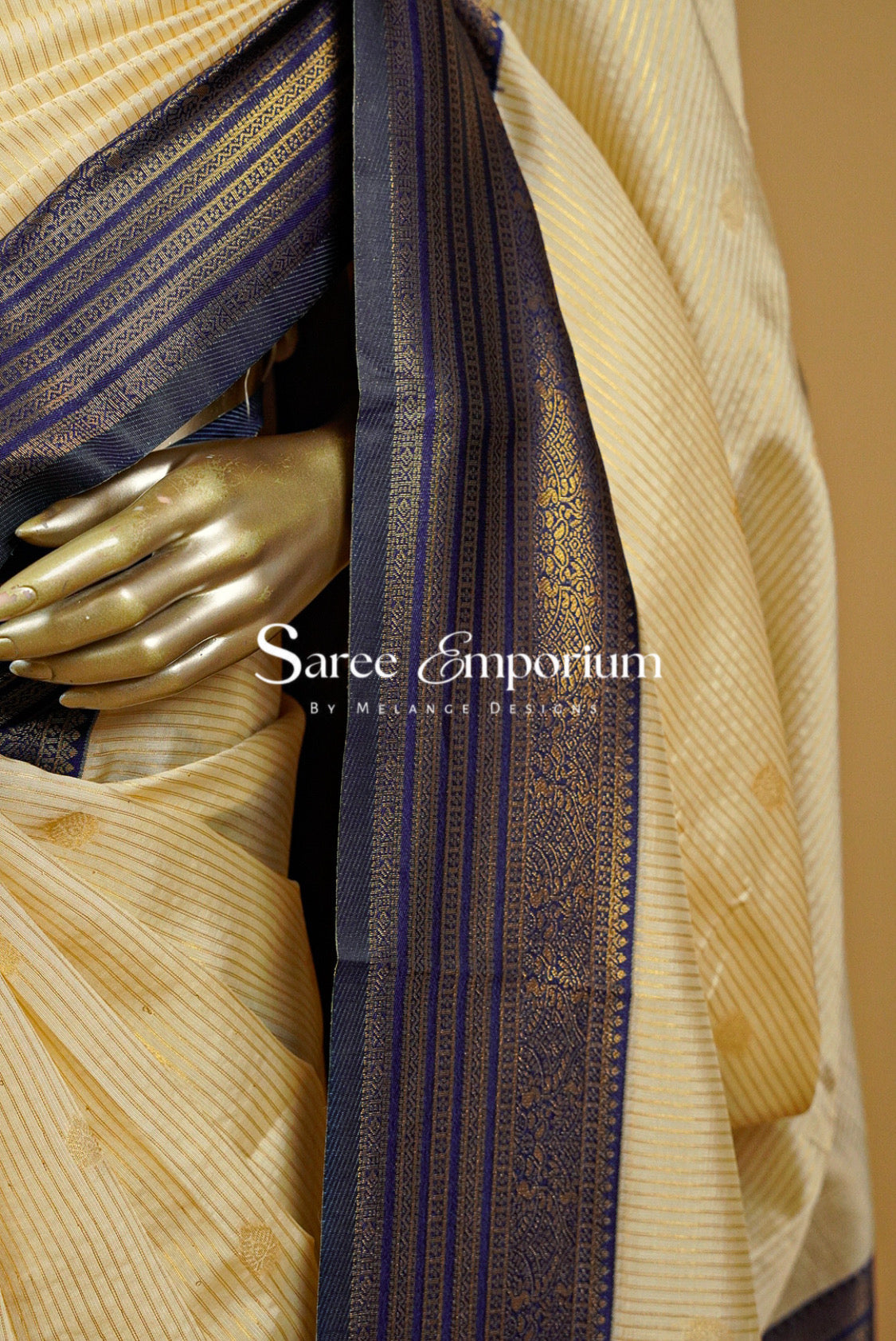 A close-up of a cream-colored silk saree with gold zari stripes and a dark navy blue border with gold motifs.