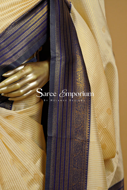 A close-up of a cream-colored silk saree with gold zari stripes and a dark navy blue border with gold motifs.