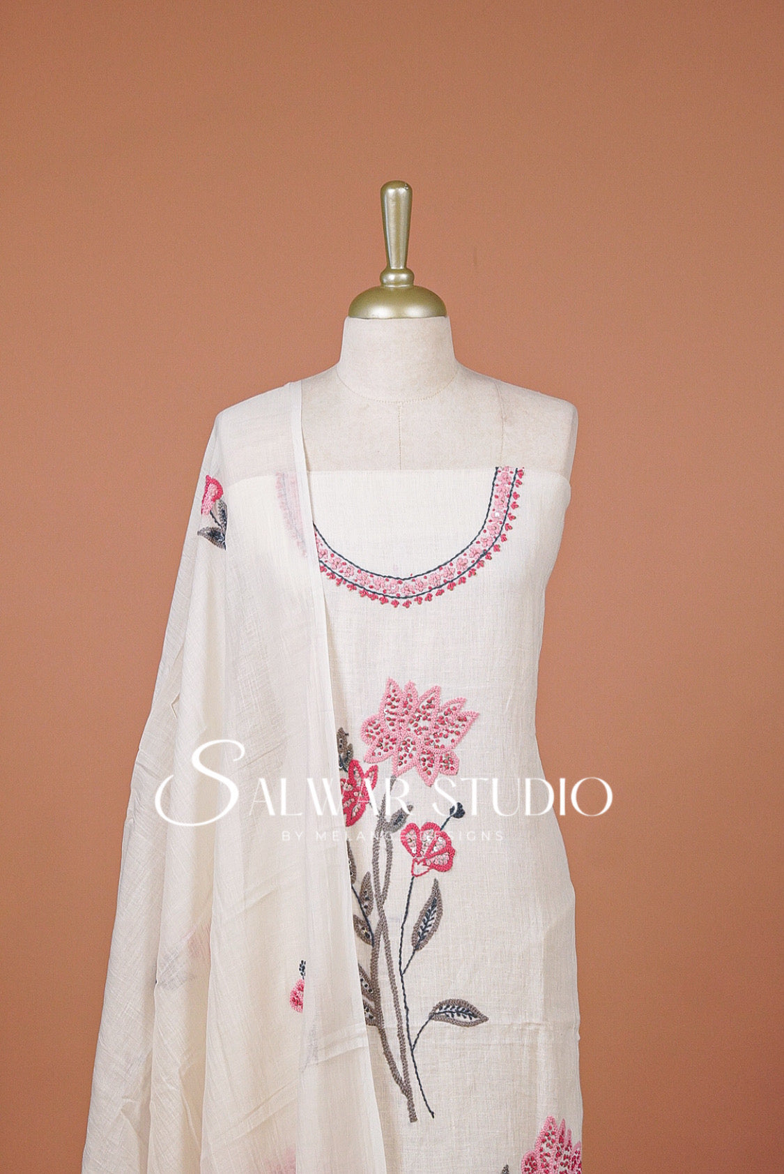 An off-white cotton fabric set displayed on a mannequin against a solid terracotta background. The set includes a kurta with pink and green floral embroidery, a matching dupatta with pink floral prints, and a small pink embroidered patch on the chest.