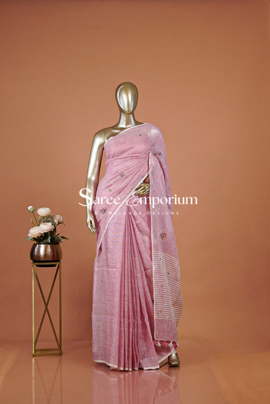 A graceful Linen Embroidery Saree in a soft blush-pink shade, featuring a beautiful natural woven texture and delicate floral embroidery scattered across the body. The understated contrast border adds a refined finish, while the striped pallu with charming tassel detailing gives the saree an elegant handcrafted appeal.