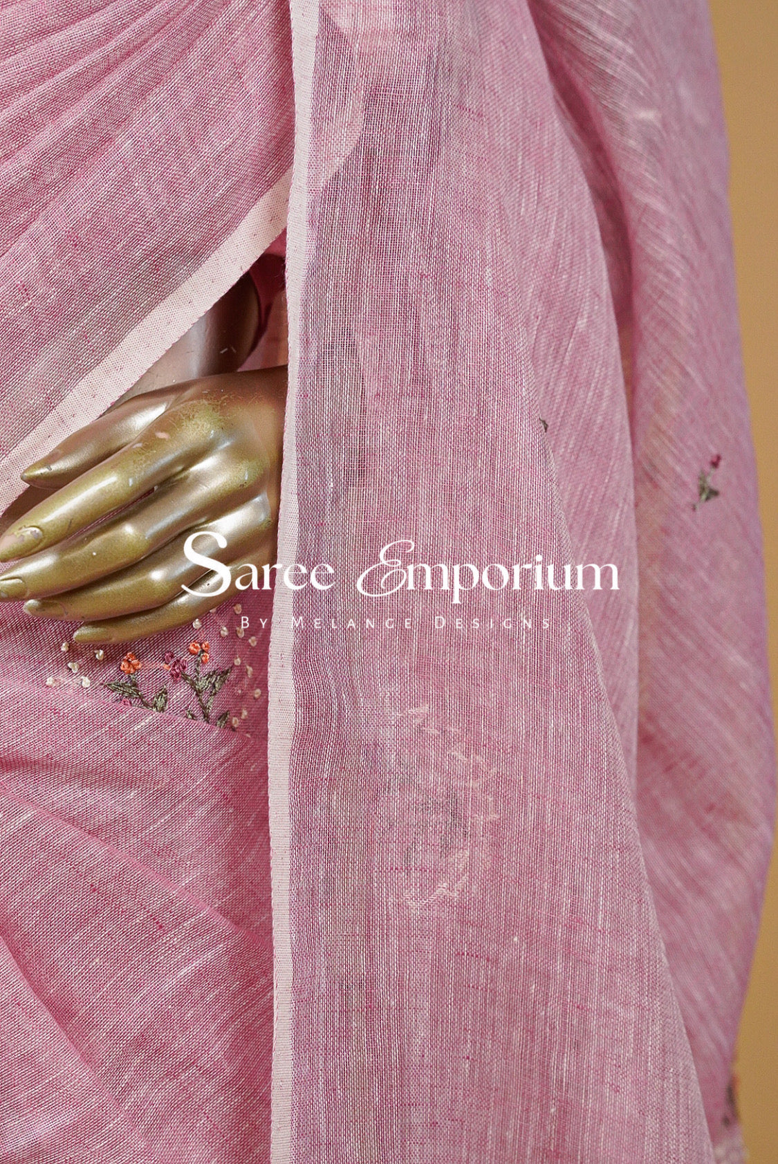 A close-up of a pink semi-sheer saree fabric draped over a gold-colored mannequin hand, featuring delicate floral embroidery .