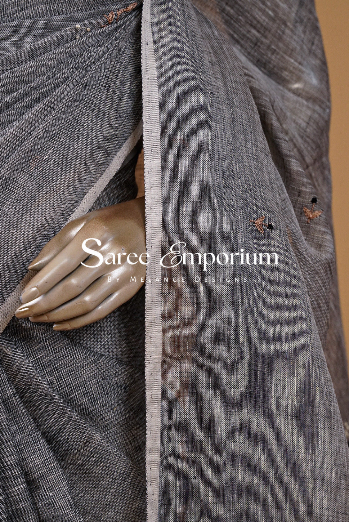 A close-up of a dark gray textured sari fabric draped elegantly, featuring a metallic gold-toned hand prop and a central vertical border. 