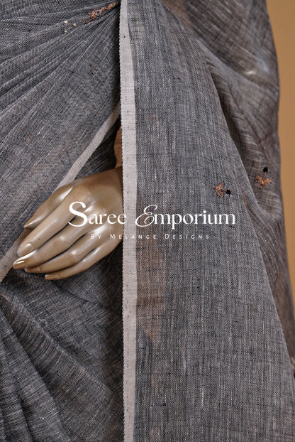 A close-up of a dark gray textured sari fabric draped elegantly, featuring a metallic gold-toned hand prop and a central vertical border. 