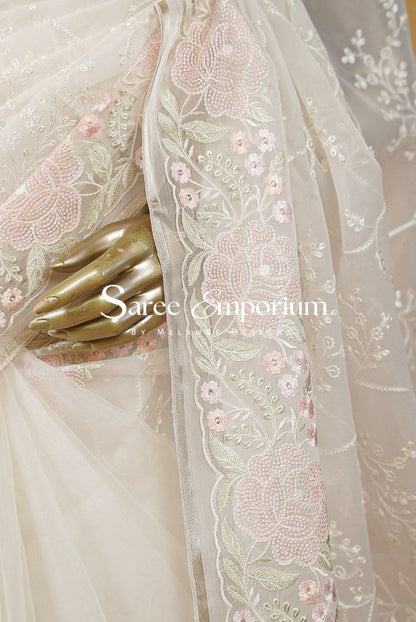 A close-up of a cream-colored saree with intricate pink and white floral embroidery on the border.