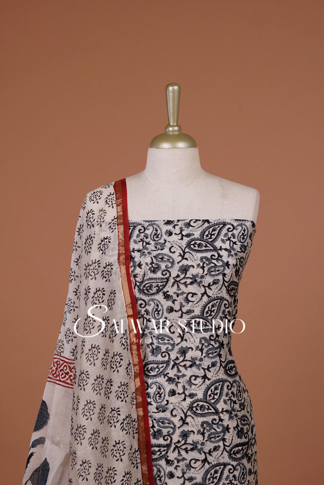 A sleeveless printed dress The garment has a black and white paisley and floral print with a red border detail along the neckline and drape.