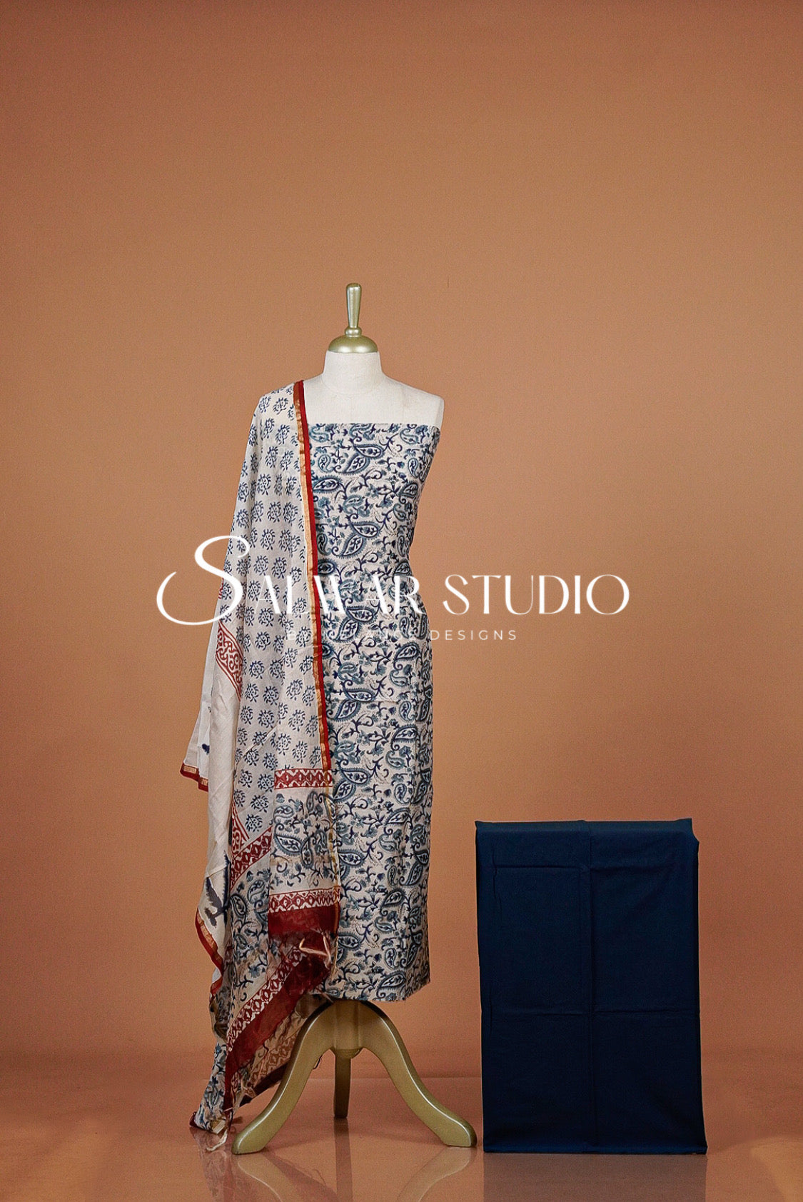 A women's three-piece salwar suit set displayed on a mannequin against a warm terracotta background. The set includes a blue and white printed kurta, a matching dupatta with red border detailing, and a navy blue folded jacket.