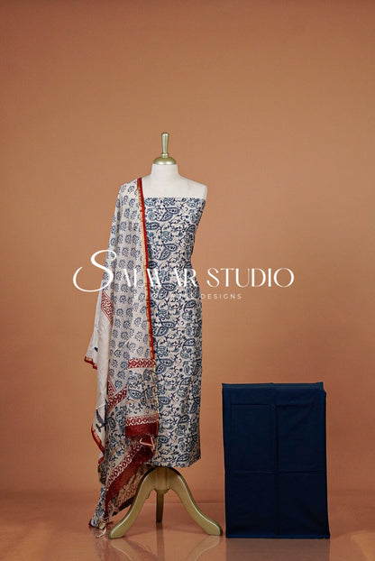A women's three-piece salwar suit set displayed on a mannequin against a warm terracotta background. The set includes a blue and white printed kurta, a matching dupatta with red border detailing, and a navy blue folded jacket.