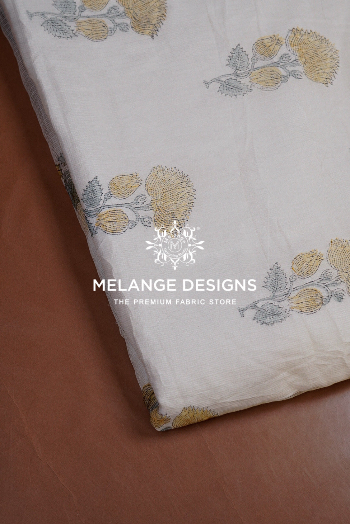 A close-up of a white Printed Cotton Cotta Fabric with a block-printed floral pattern in muted yellow and gray, draped diagonally over a brown leather surface. 