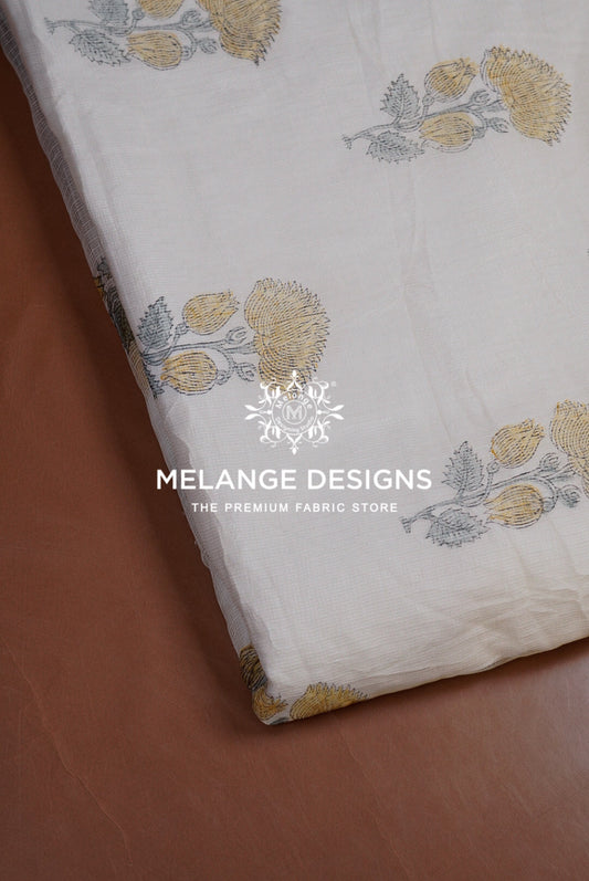 A close-up of a white Printed Cotton Cotta Fabric with a block-printed floral pattern in muted yellow and gray, draped diagonally over a brown leather surface. 