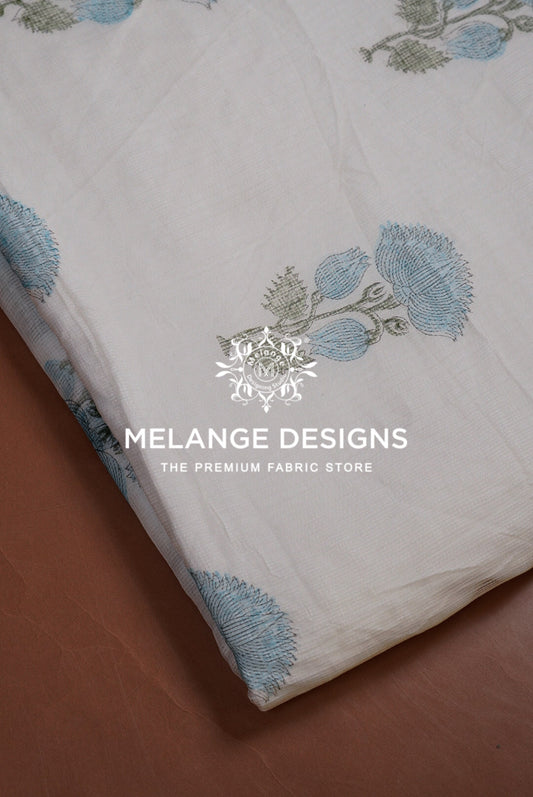 A close-up of a white Printed Cotton Cotta Fabric with light blue and gray floral block prints, folded diagonally on a brown surface. 