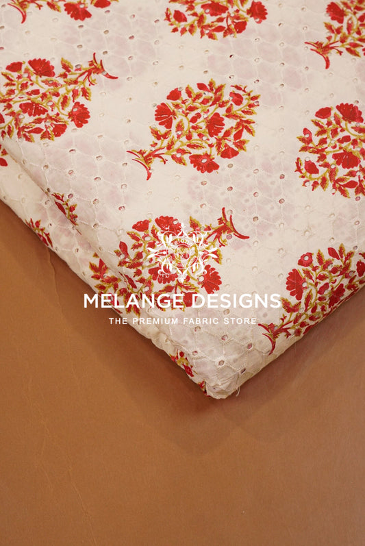 A folded piece of white eyelet lace Printed Cotton Hacoba Fabric with a red and orange floral pattern, displayed on a brown surface .