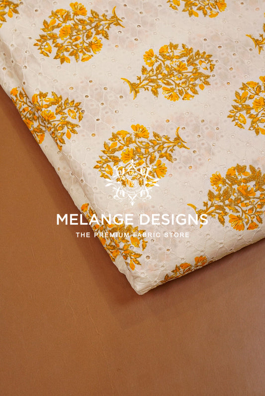 A folded piece of off-white cotton Printed Cotton Hacoba Fabric with a yellow floral print and eyelet embroidery, displayed on a brown surface .