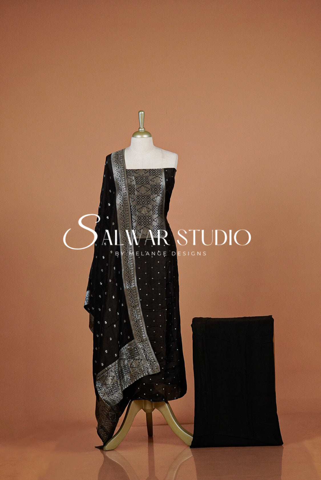 A black salwar suit set with a matching black fabric bag or accessory beside it. The set includes a draped top and dupatta with silver mirror-work border detailing.
