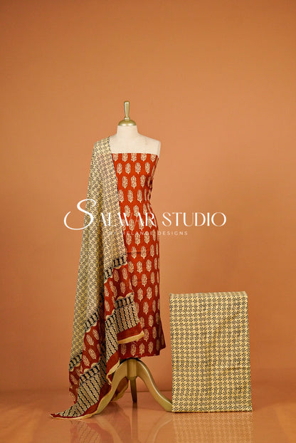 A women's salwar suit set  The set includes a rust-orange salwar with cream and gold motifs, a matching beige dupatta with black and gold geometric patterns, and a folded matching fabric piece.