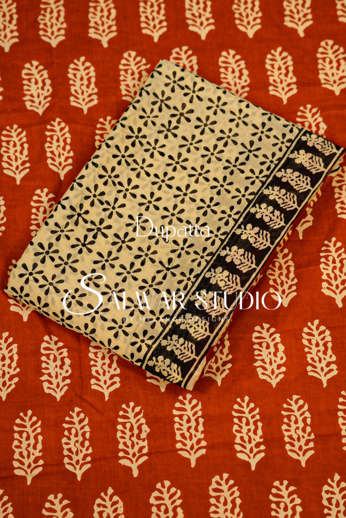 A folded block-printed cotton fabric piece with a beige base and black floral motifs, displayed diagonally on a red-orange fabric background with cream leaf prints. 