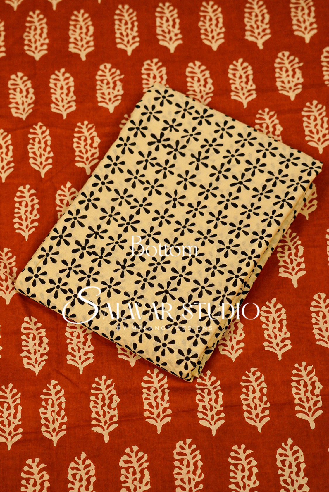 A folded beige fabric item with a black floral block print pattern, placed diagonally on a rust-red fabric background with cream leaf motifs. 