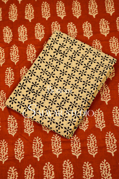 A folded beige fabric item with a black floral block print pattern, placed diagonally on a rust-red fabric background with cream leaf motifs. 