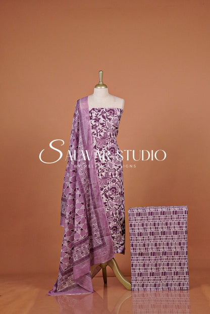 A purple and white printed traditional dress set featuring a matching patterned dupatta draped over the shoulder and a folded fabric swatch beside .