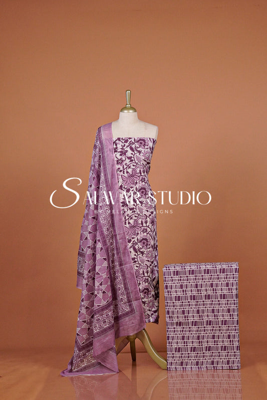 A purple and white printed traditional dress set featuring a matching patterned dupatta draped over the shoulder and a folded fabric swatch beside .