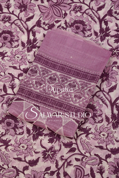 A folded dusty purple dupatta with intricate floral and paisley patterns, on top of a matching patterned fabric. 