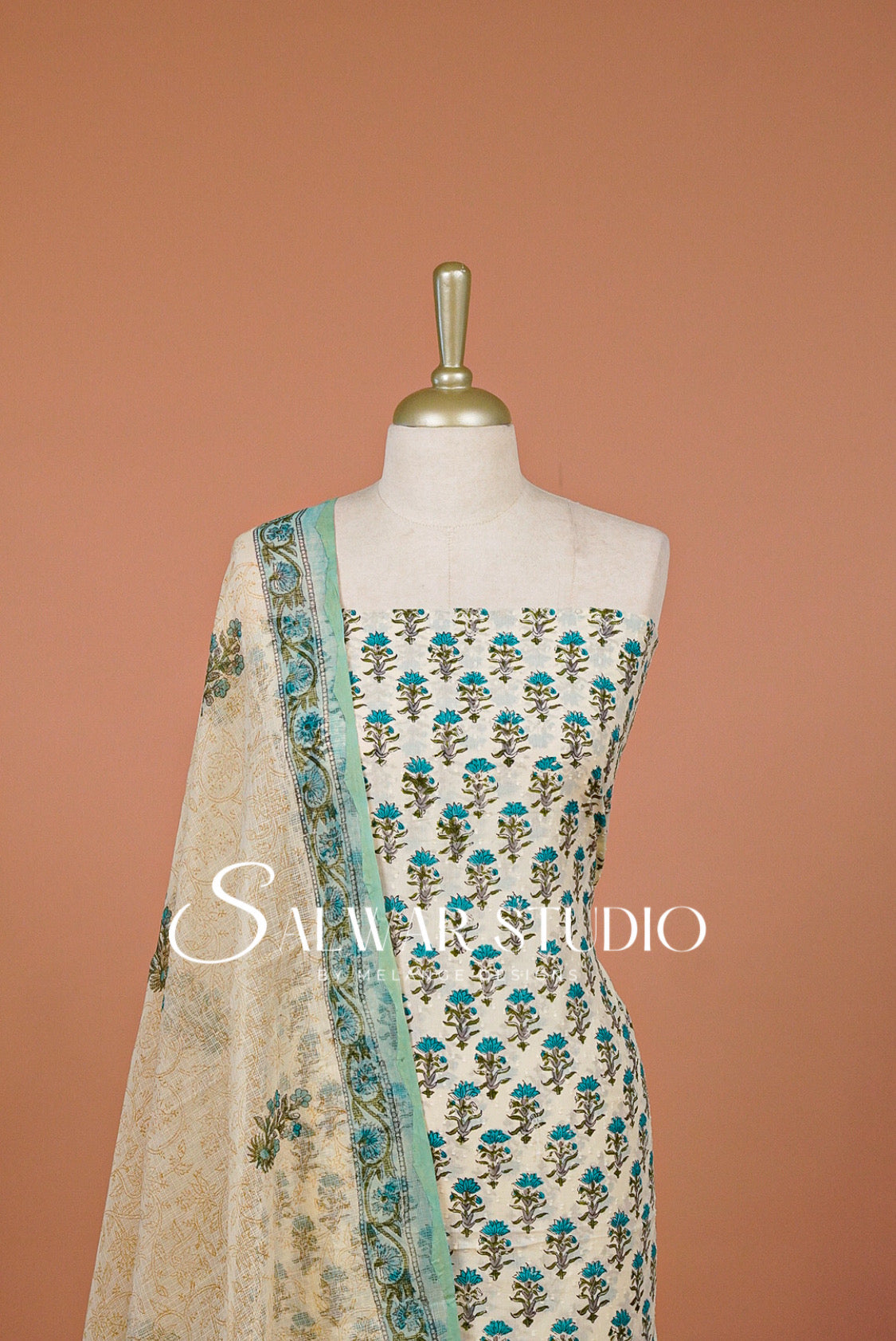 A cream-colored Indian ethnic salwar suit set .The set includes a printed kurta with teal and green floral motifs, a matching printed dupatta, and a light blue border on the kurta's edge.