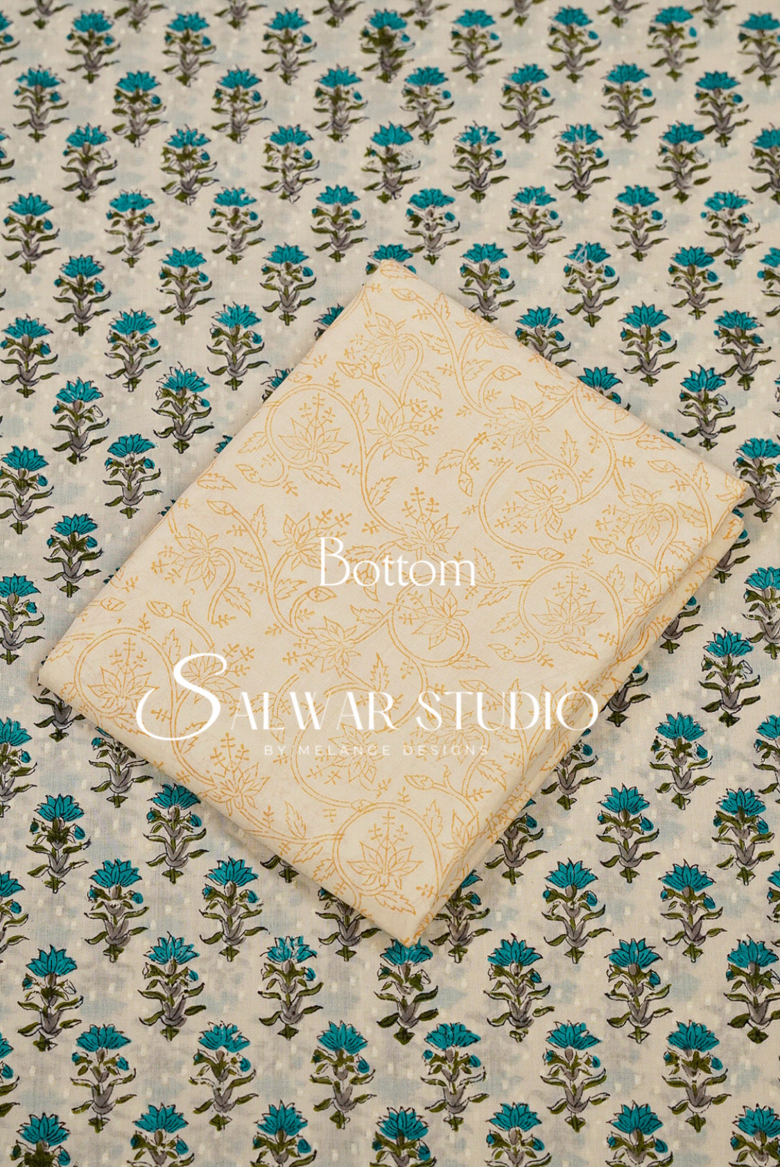 A folded cream-colored fabric piece with a subtle gold floral pattern, placed diagonally on top of a larger matching fabric piece featuring a repeating teal and brown block print on a light beige .