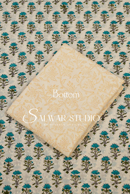 A folded cream-colored fabric piece with a subtle gold floral pattern, placed diagonally on top of a larger matching fabric piece featuring a repeating teal and brown block print on a light beige .
