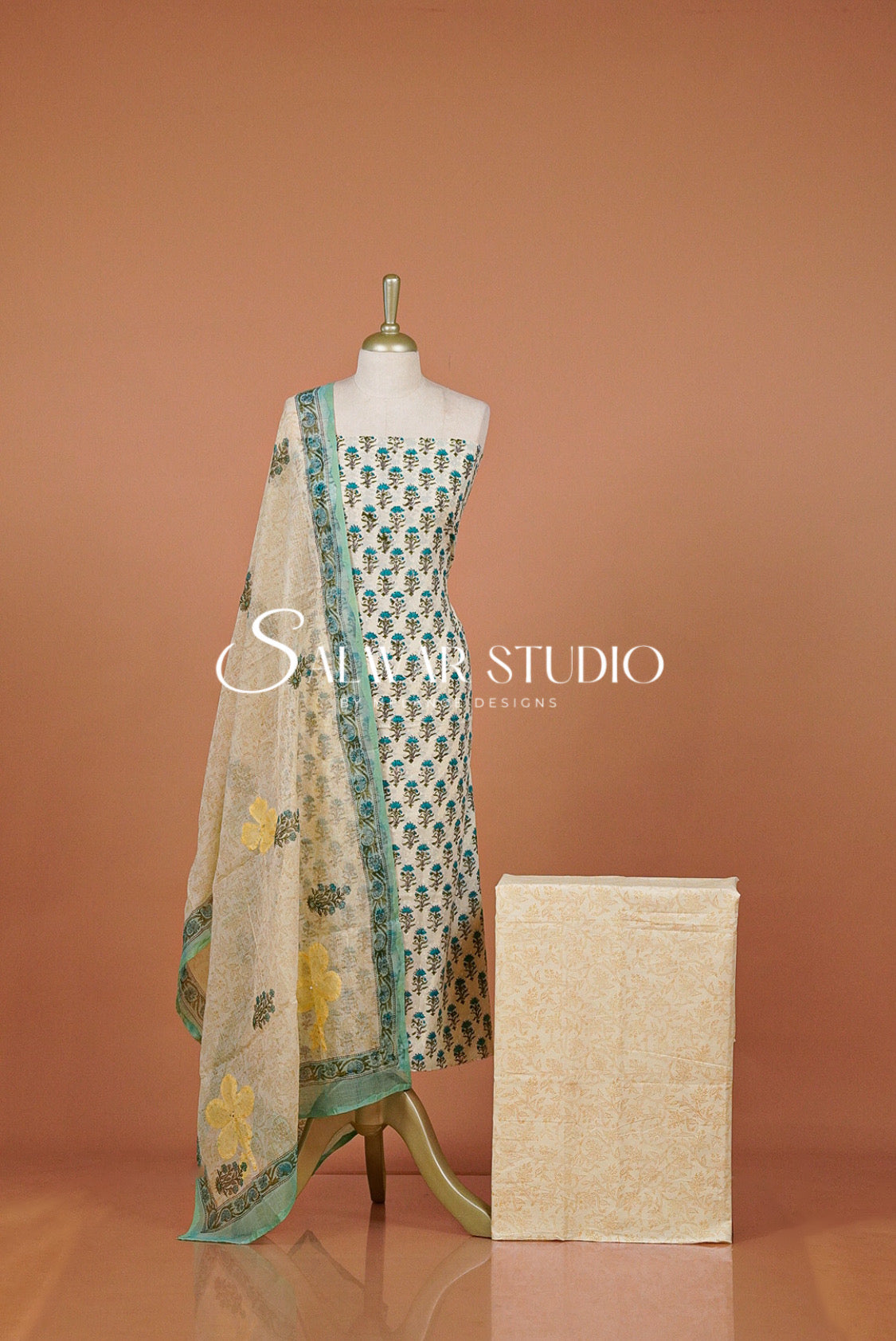 A women's three-piece salwar suit set. The set includes a light beige printed kurta with teal and yellow floral motifs, a matching teal border dupatta, and a folded beige printed fabric piece beside it.