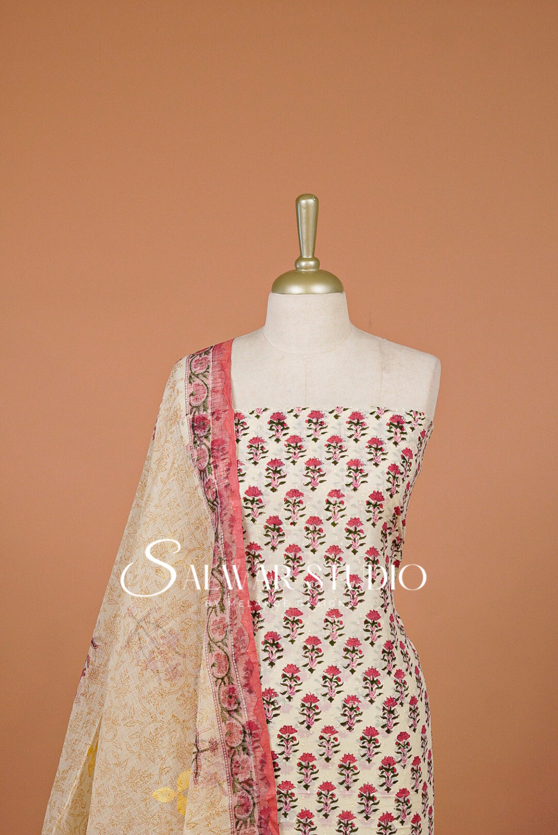 A cream-colored three-piece salwar kameez suit set .The set includes a printed kameez top with pink and green floral motifs, a beige dupatta with a pink border, and a matching beige printed bottom fabric.