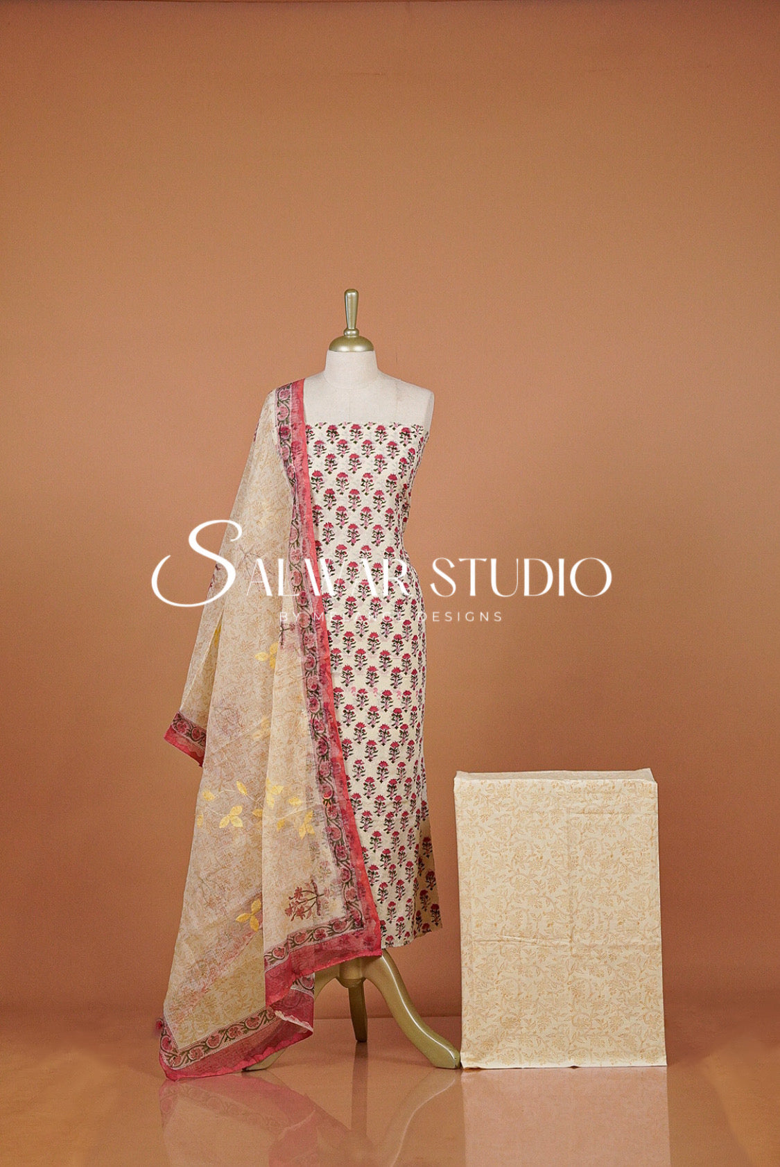 A traditional Indian salwar suit set . The set includes a beige printed salwar kameez with red and gold floral motifs, a matching beige dupatta with red borders, and a folded beige fabric piece.