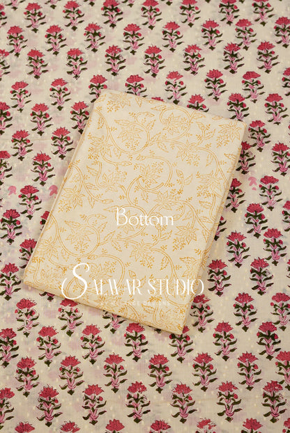 A folded cream-colored fabric piece with gold floral patterns lies on top of a larger matching fabric sheet featuring a repeating pink and green block print on a cream background. 